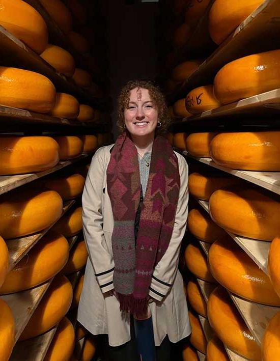 A Converse student stands smiling between massive wheels of cheese at a cheese museum in Amsterdam.